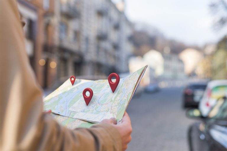 Pessoa segurando um mapa com ícones de localização em vermelho sobrepostos, em uma rua urbana com prédios e carros ao fundo desfocado.
