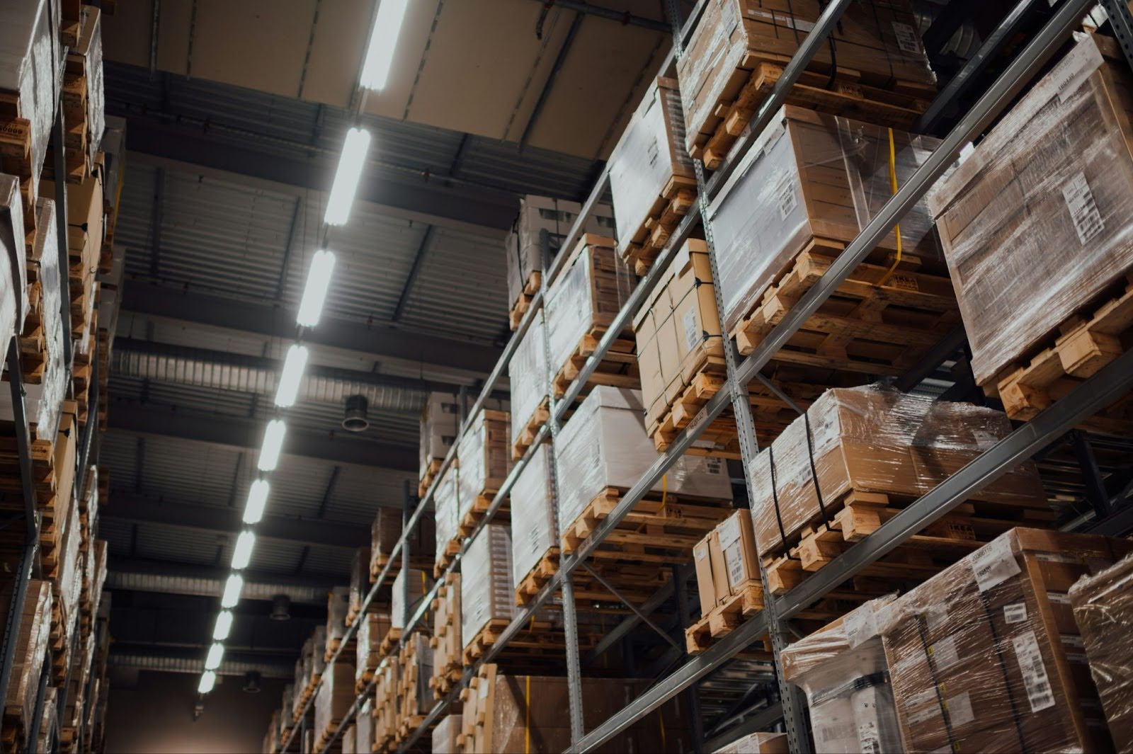 Interior de um armazém logístico com prateleiras industriais altas repletas de caixas em paletes, iluminado por fileiras de luzes LED no teto.